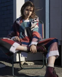 Model seated outdoors in a colorful patchwork knitted sweater and matching trousers with dark boots.