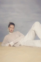 Model lying on sandy dunes wearing a white textured turtleneck and trousers against an overcast sky.