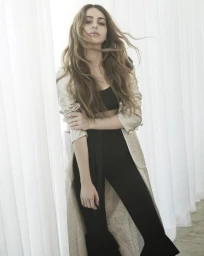 Woman with long flowing hair wearing a long coat and black outfit against white curtains
