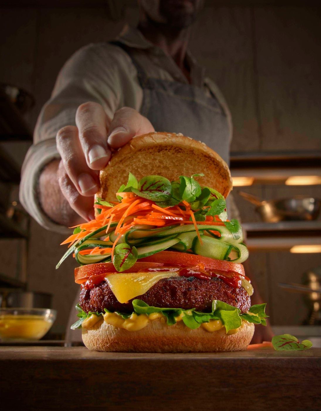 Chef presenting a freshly made veggie burger with layers of fresh ingredients, including lettuce, tomato, and shredded carrots.