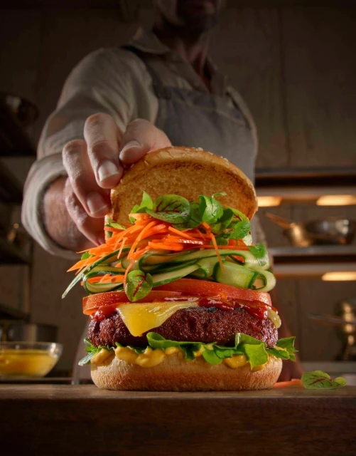 Chef presenting a freshly made veggie burger with layers of fresh ingredients, including lettuce, tomato, and shredded carrots.