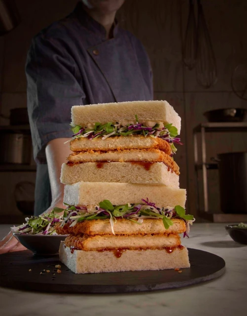 Stacked katsu sando towers with salad, prepared by chef in background. Close-up food photography captures the vibrant colors and textures.