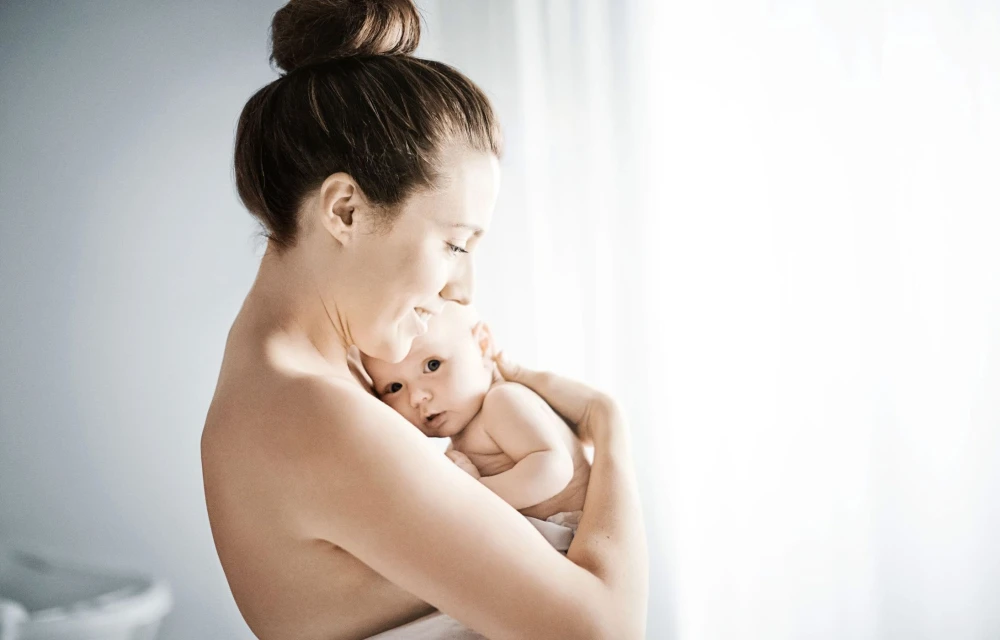A mother lovingly holds her baby in a soft, bright indoor setting with a gentle expression of joy and tenderness.