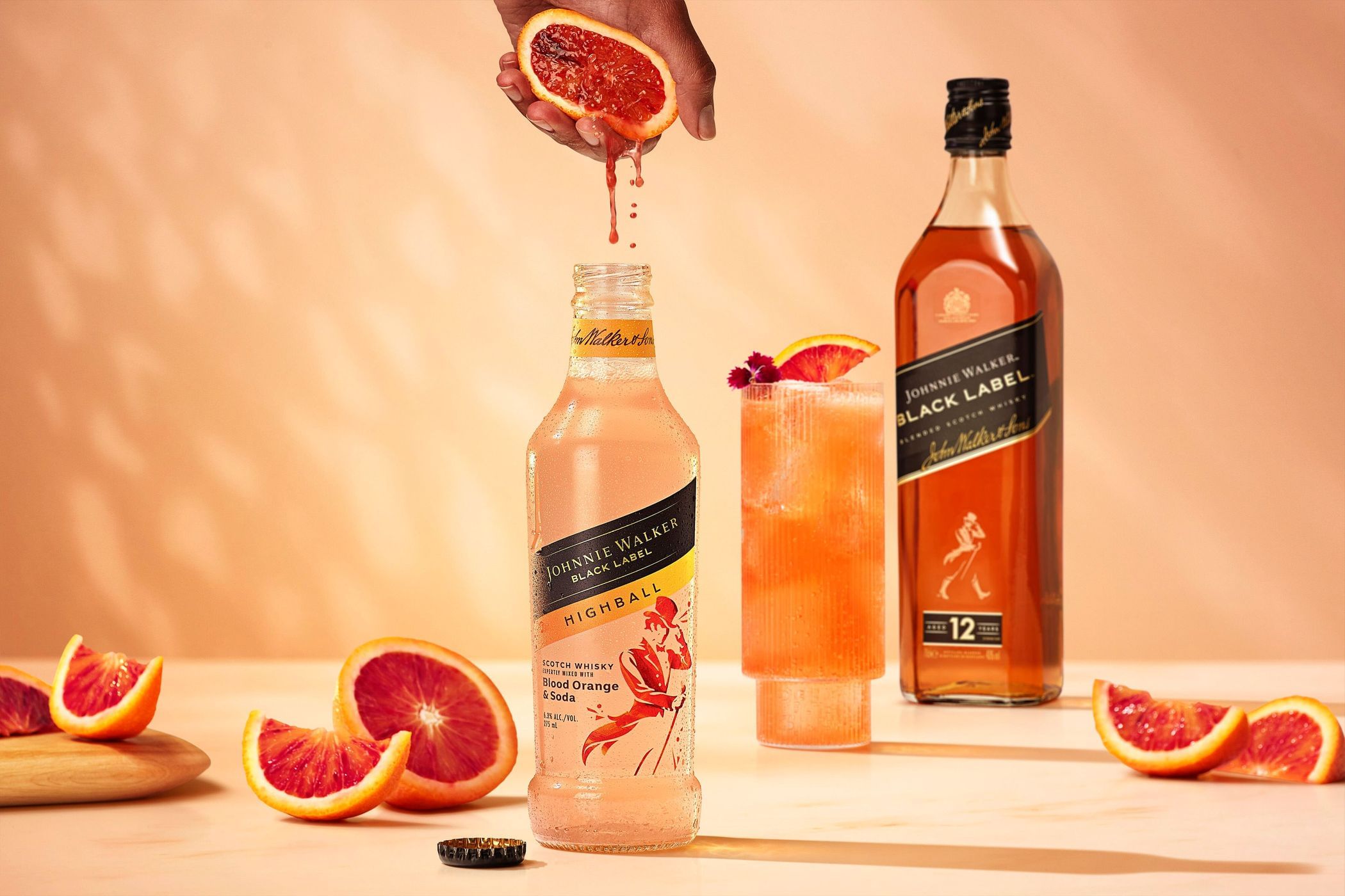 Blood orange juice pouring into Johnnie Walker Highball cocktail, with bottle, whisky, and orange slices against peach background.