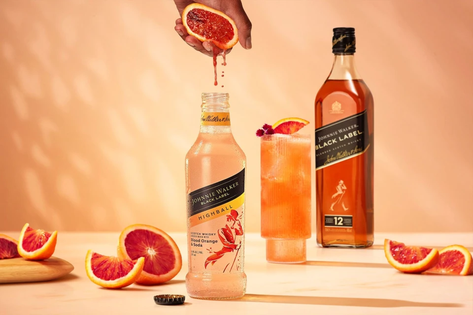 Blood orange juice pouring into Johnnie Walker Highball cocktail, with bottle, whisky, and orange slices against peach background.