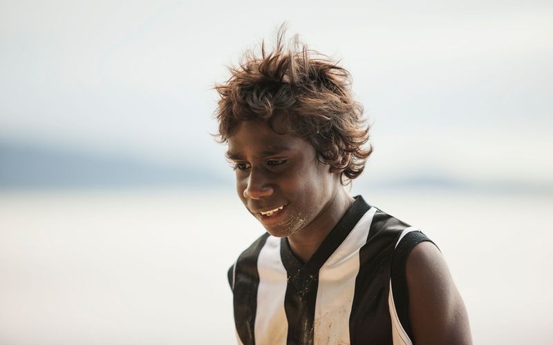 Portrait of an Indigenous Australian child with curly hair, smiling gently, wearing a striped sports jersey outdoors on a sandy shore.