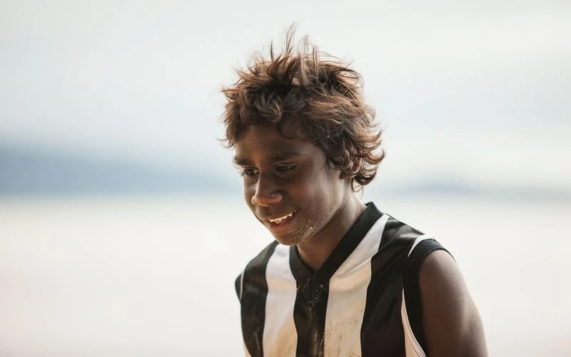 Portrait of an Indigenous Australian child with curly hair, smiling gently, wearing a striped sports jersey outdoors on a sandy shore.
