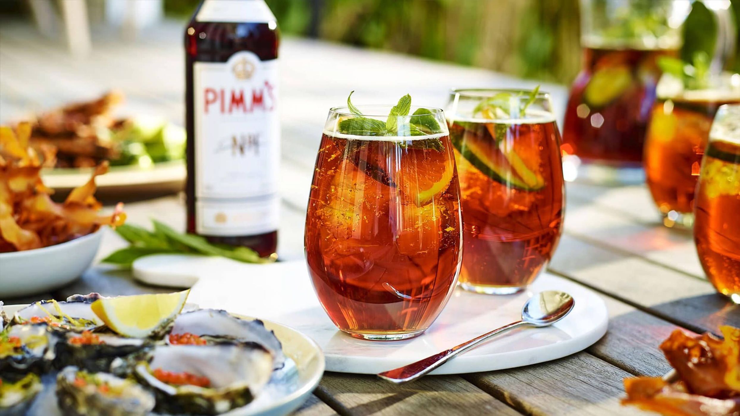 Close-up of two Pimm's cocktails garnished with mint and fruit, oysters, and a bottle of Pimm's on a white wooden table.