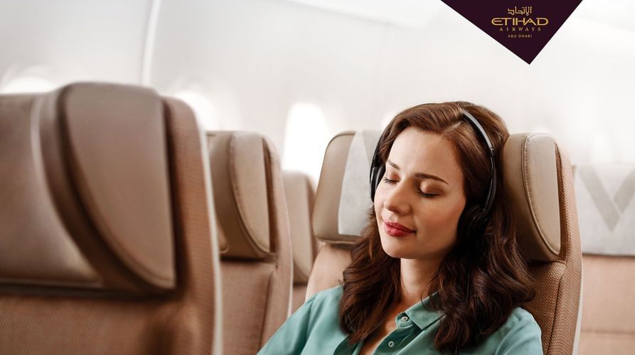 A woman with headphones on a plane relaxes in her seat with her eyes closed, embodying comfortable air travel with Etihad Airways.