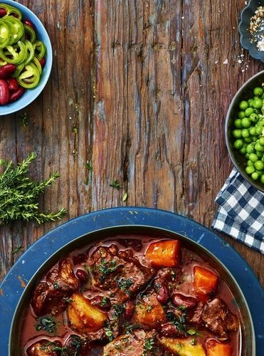 Overhead shot of a vibrant stew featuring meat, potatoes, and beans, set on a wooden table next to bowls of peppers and peas.