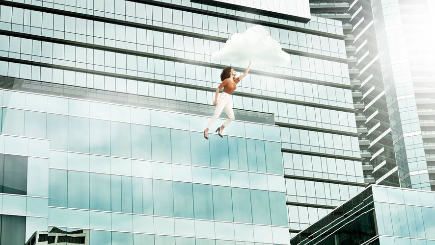 Woman in white pants and heels floats holding a cloud, with high-rise glass buildings in the background, under bright, natural lighting.