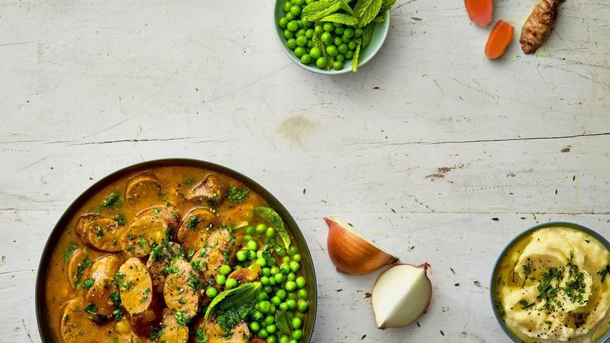 Overhead shot of a hearty sausage stew in a dark bowl, garnished with herbs and peas, and paired with carrots, onions, and mashed potatoes.