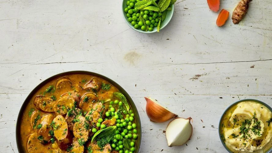 Overhead shot of a hearty sausage stew in a dark bowl, garnished with herbs and peas, and paired with carrots, onions, and mashed potatoes.