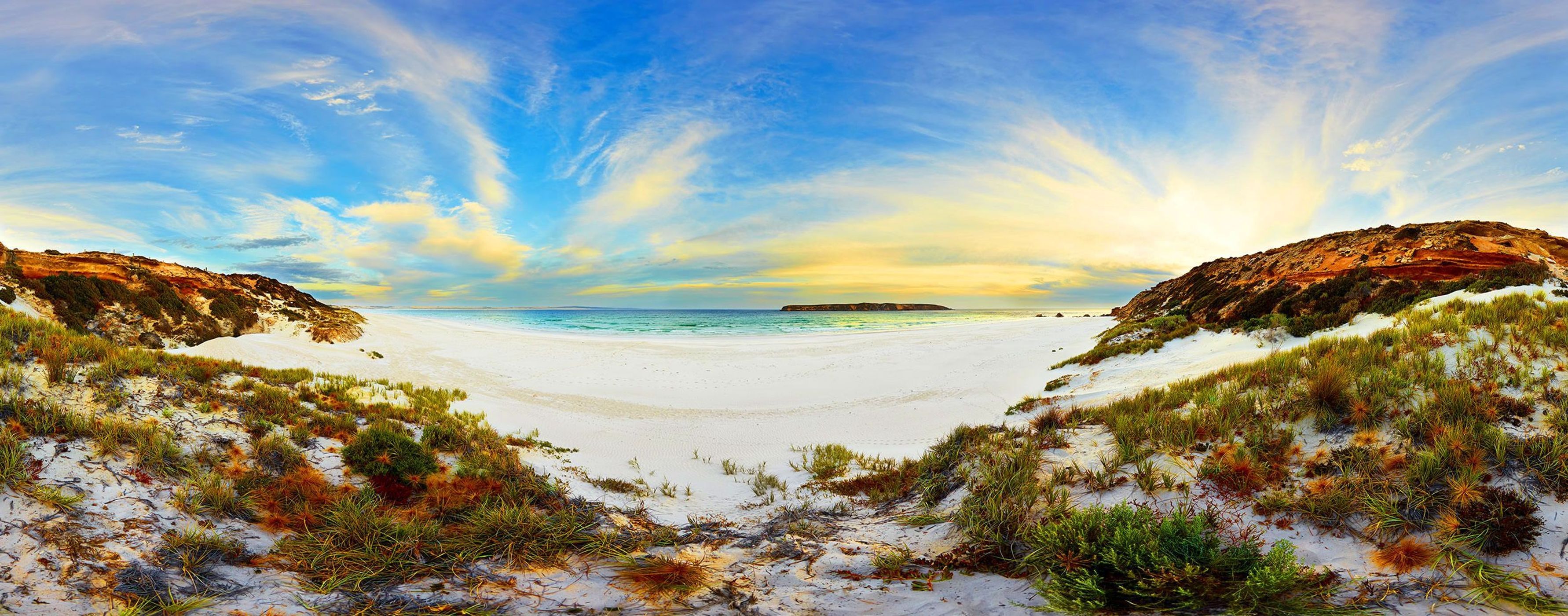 Panoramic view of a white sand beach leading to a turquoise ocean, flanked by grassy dunes and hills under a blue and yellow sky.