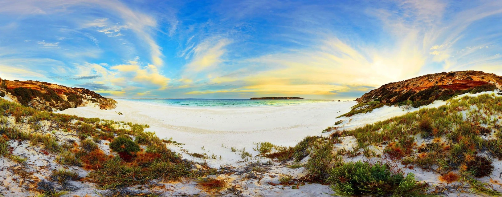 Panoramic view of a white sand beach leading to a turquoise ocean, flanked by grassy dunes and hills under a blue and yellow sky.