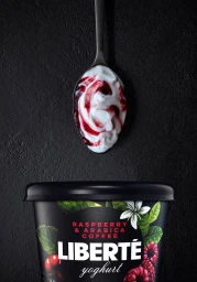 A black spoon filled with white yogurt swirled with red raspberry jam held above a black Liberte yogurt container.