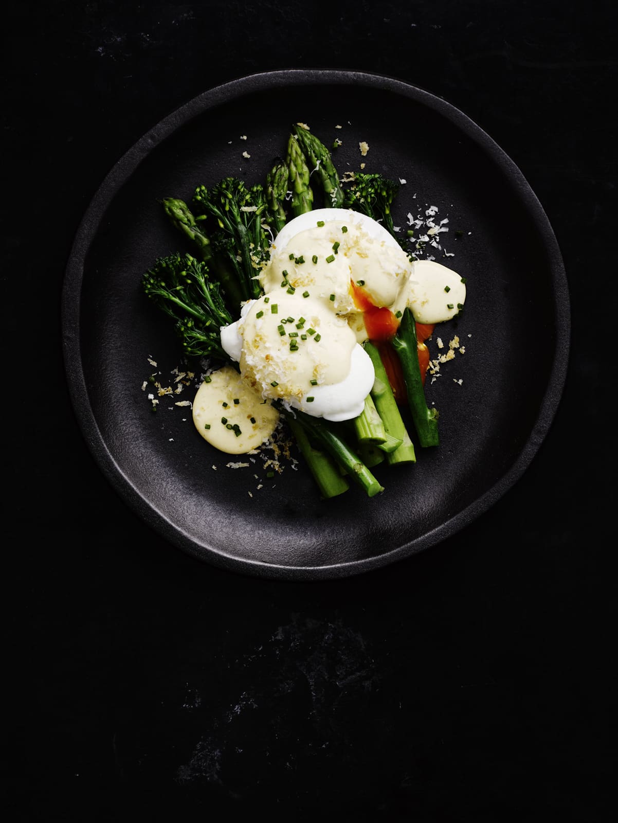 Overhead view of a black plate with two poached eggs on asparagus and broccolini, topped with sauce and chives, food photography.