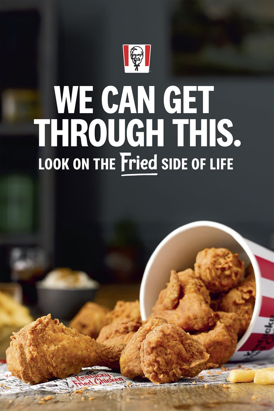 Fried chicken pieces spill out of a bucket onto a table with a dark background, promoting KFC's slogan about enjoying the fried side of life.