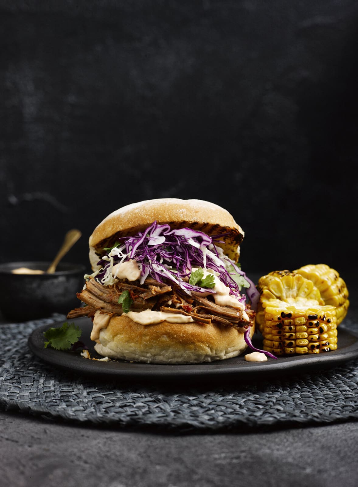 Burger with pulled pork and purple cabbage on a dark plate, garnished with greens, served with grilled corn and a dipping sauce.