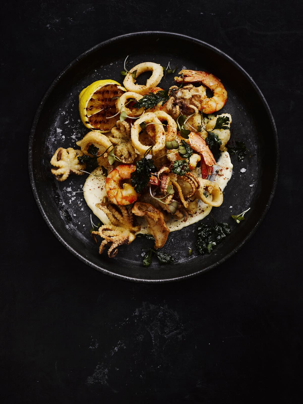Overhead shot of seafood including shrimp, calamari, octopus, and lemon on a black plate against a dark background.