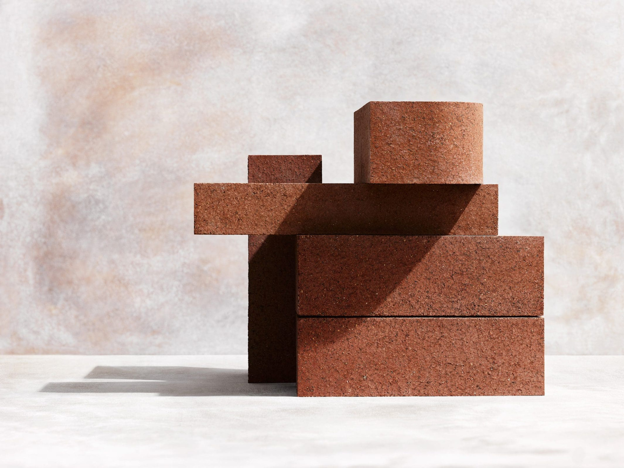 Still life of stacked geometric shapes in red-brown tones against a neutral textured background, with a play of light and shadow.