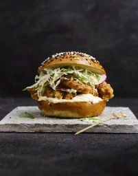 Close-up food photography of a gourmet fried chicken burger with shredded cabbage slaw on a stone tile by Jon Bader.