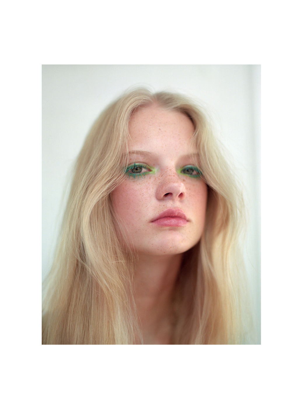 Portrait of a young woman with long blond hair and green makeup around her eyes, posing in front of a plain white wall.