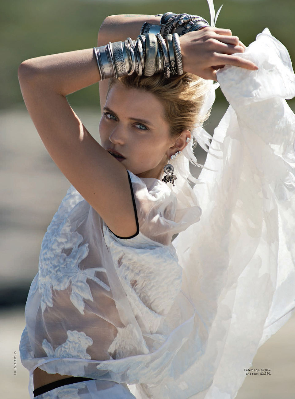 Fashion shot of a blonde woman wearing silver bracelets and a white sheer top, posing outdoors in natural light, editorial photography.