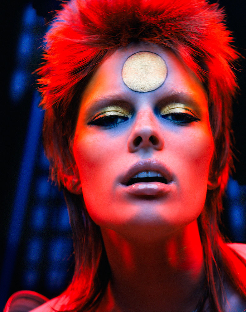 Close-up of a woman with distinctive makeup, gold eyeshadow and a gold circle on her forehead, set against a blurred background.