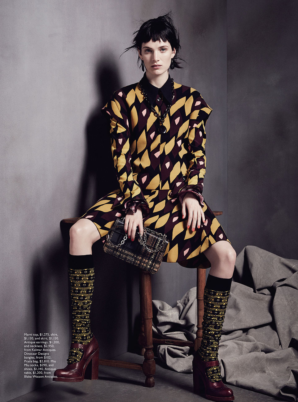 Fashion editorial shot of a woman sitting on a wooden stool, wearing a patterned dress, knee-high socks, and platform heels, with a gray backdrop.