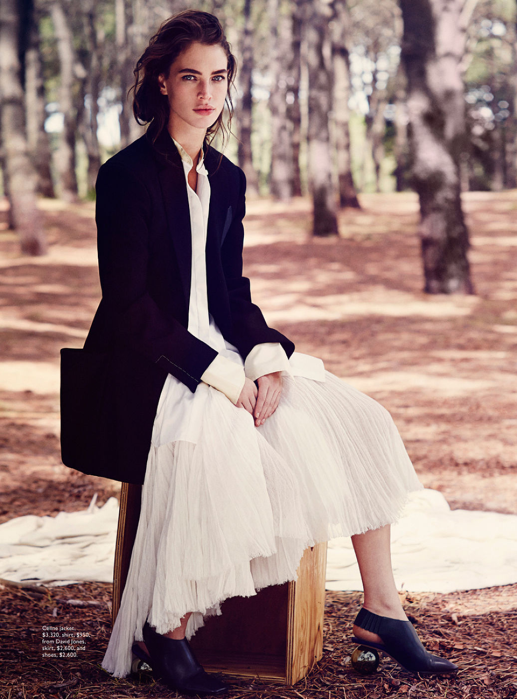 A woman sits on a wood box in a forest wearing a white pleated dress and black jacket for a fashion shot with trees behind her.