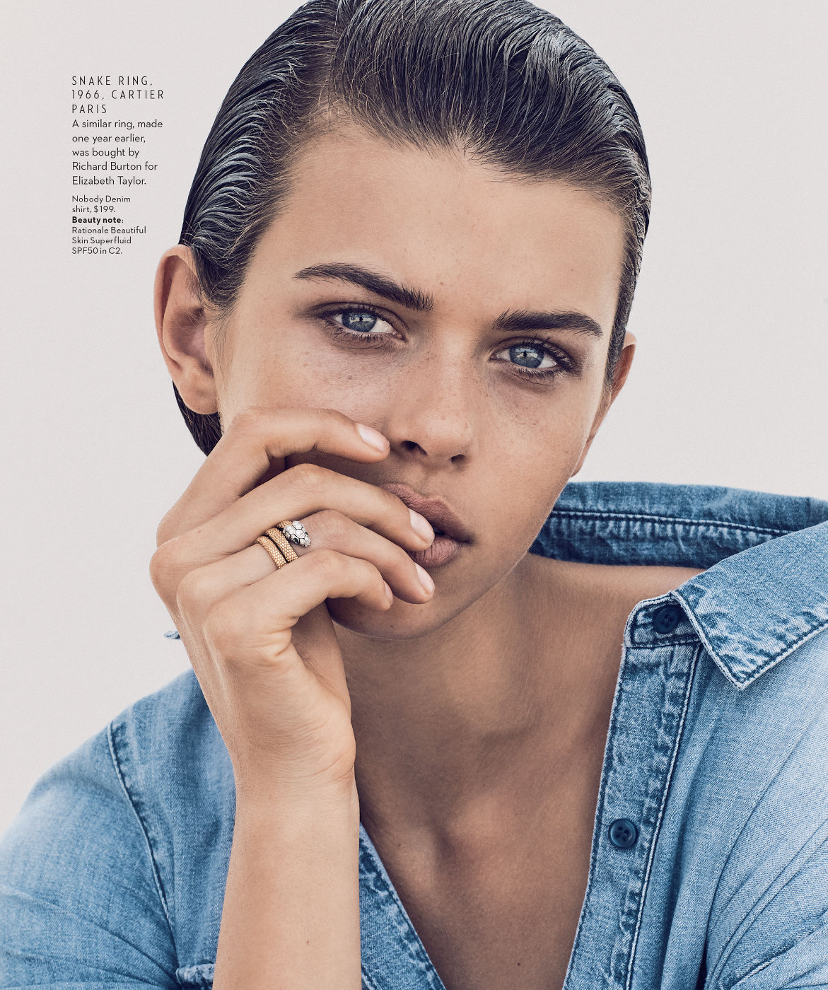 Close-up of a woman with slicked-back hair, wearing a blue denim shirt and gold rings while posing indoors.