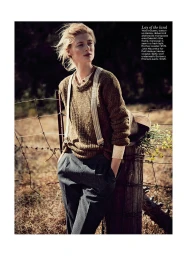 Woman in a brown knitted sweater and grey trousers with suspenders leaning against a wooden fence post in a rural field