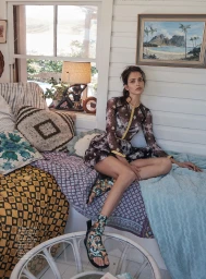 Woman seated on a daybed inside a rustic room with patterned textiles and a window overlooking a landscape painting