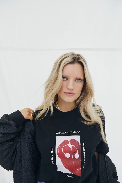 A young woman with blonde hair poses in a black Camilla and Marc sweatshirt with a hand design and text against a white backdrop.
