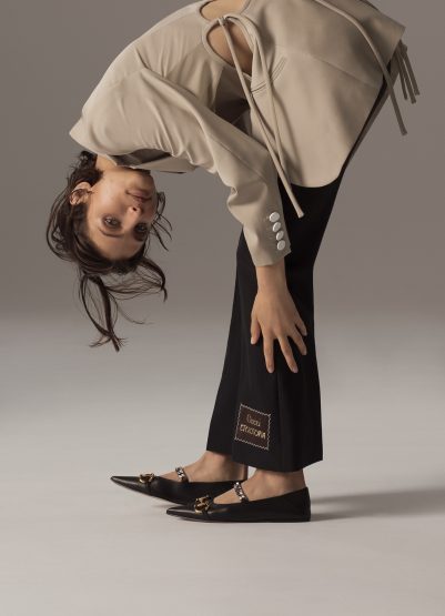A stylish young woman in a cream-colored jacket and black pants poses in a studio, bending over with dark hair, and wearing black flats.
