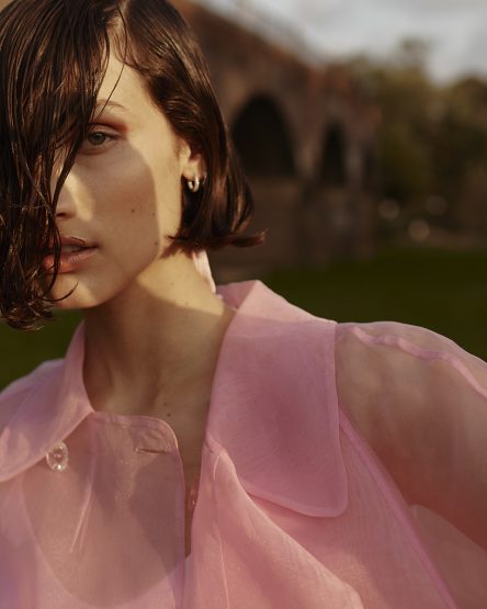 Close-up portrait of a young adult woman with dark hair, wearing a pink shirt, standing outdoors in front of an old arched bridge.