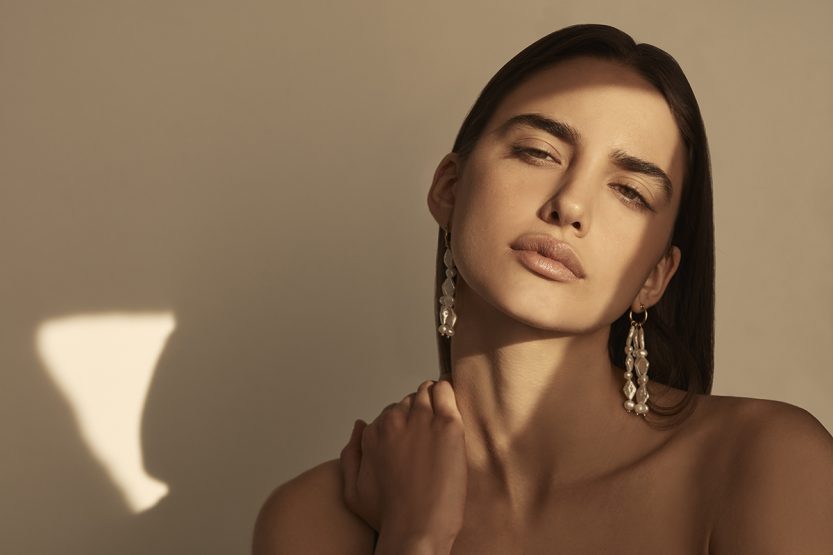 Close-up studio portrait of a young adult woman with dark hair, wearing statement earrings, and posing with her hand on her shoulder.