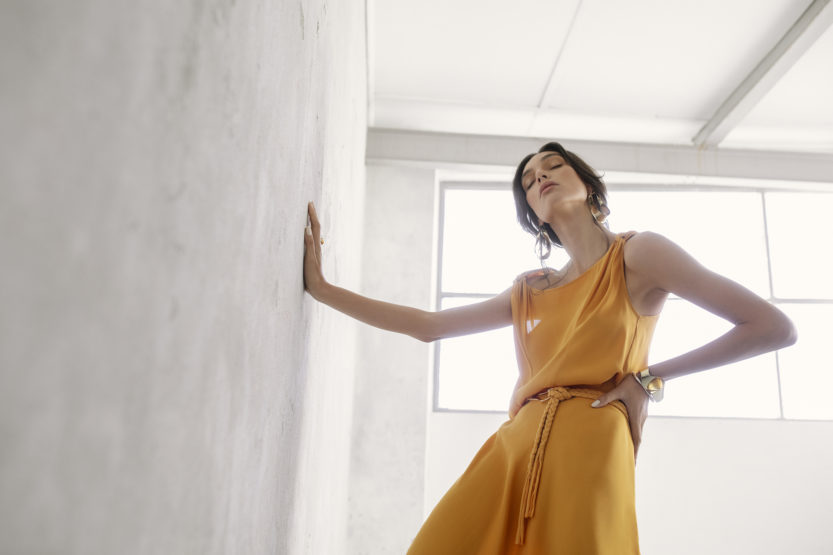 Adult woman in a yellow dress poses next to a wall in an indoor setting with natural light from the window. 