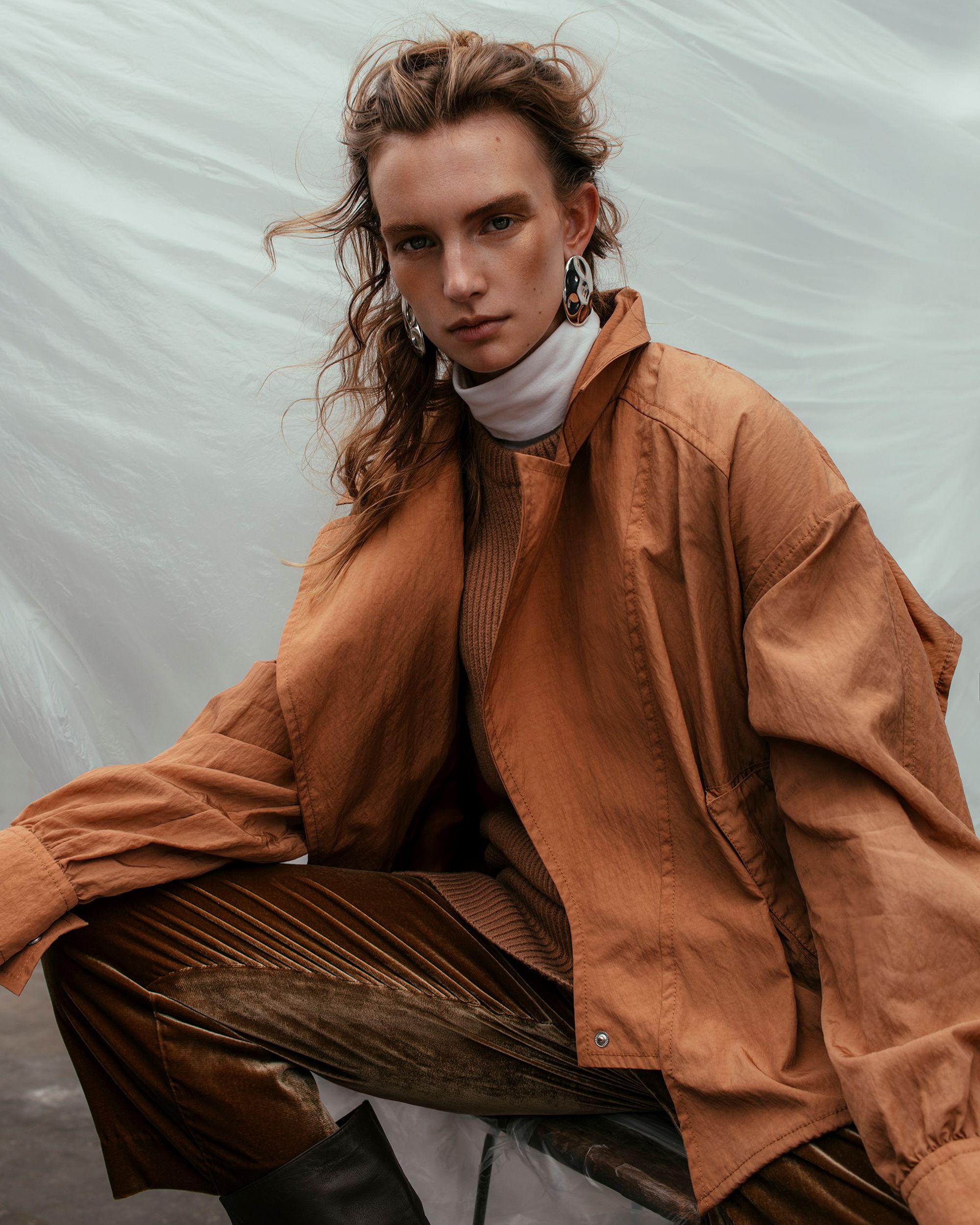 A fashionable adult female poses in an outdoor setting, wearing a tan jacket, sweater, and velvet pants, along with large earrings.