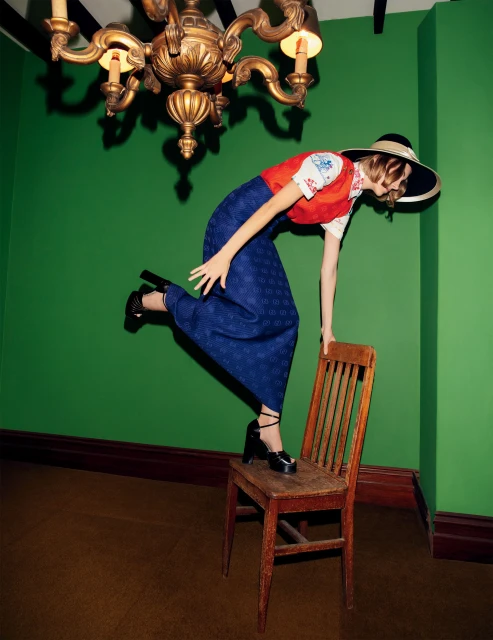 A young woman in a wide-brimmed hat and vintage-style clothing strikes a pose on a wooden chair against a green wall indoors.