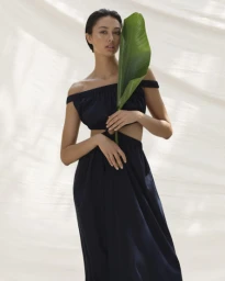 Model in off-the-shoulder dark dress holding large green tropical leaf against textured white fabric background