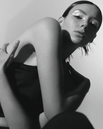 Close-up monochrome portrait of a woman with metallic eyeshadow and dark hair, posing against a white studio wall.