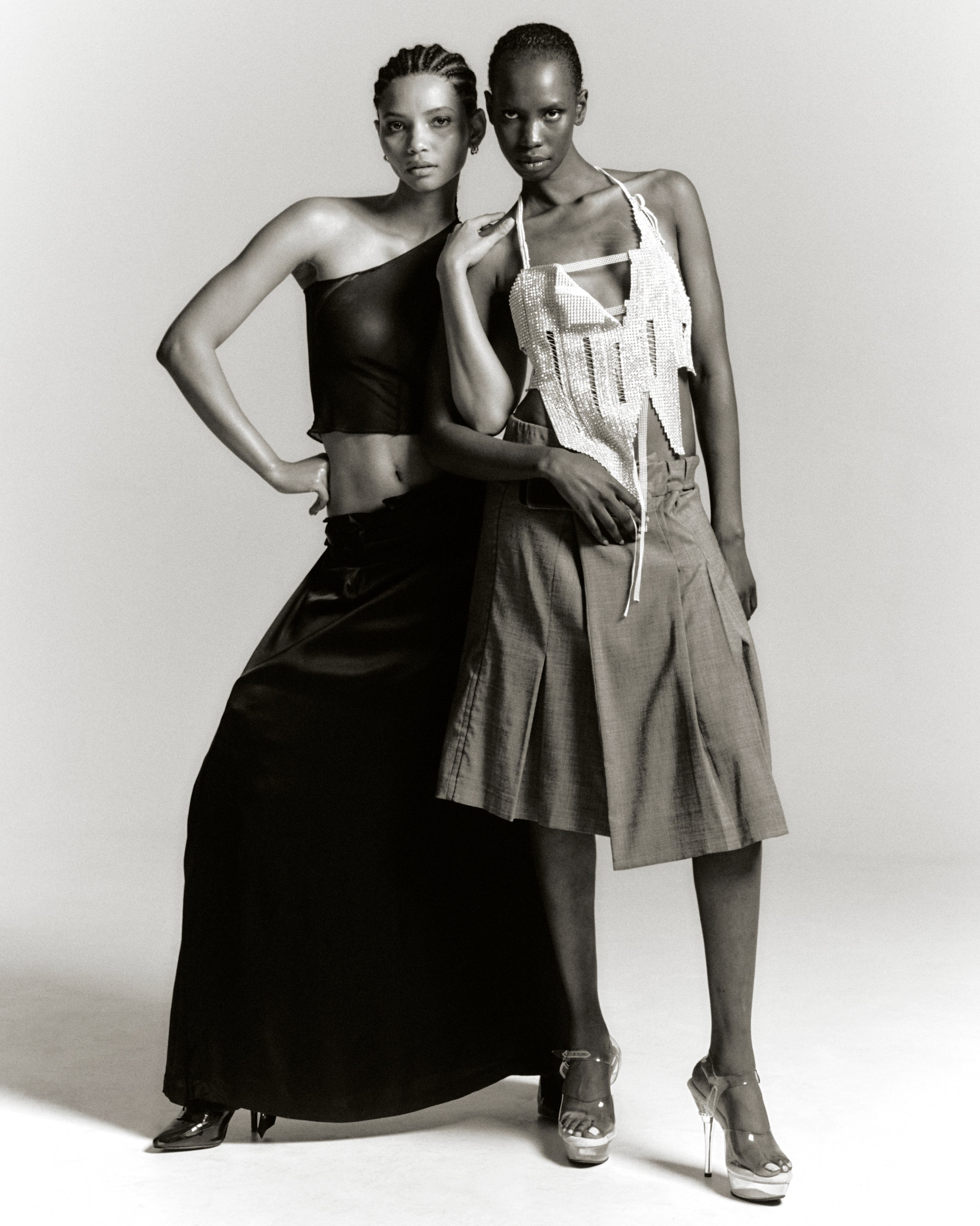 Two fashion models pose in a photo studio. One wears a black top and long black skirt; the other, a light-colored top, skirt, and platform shoes.