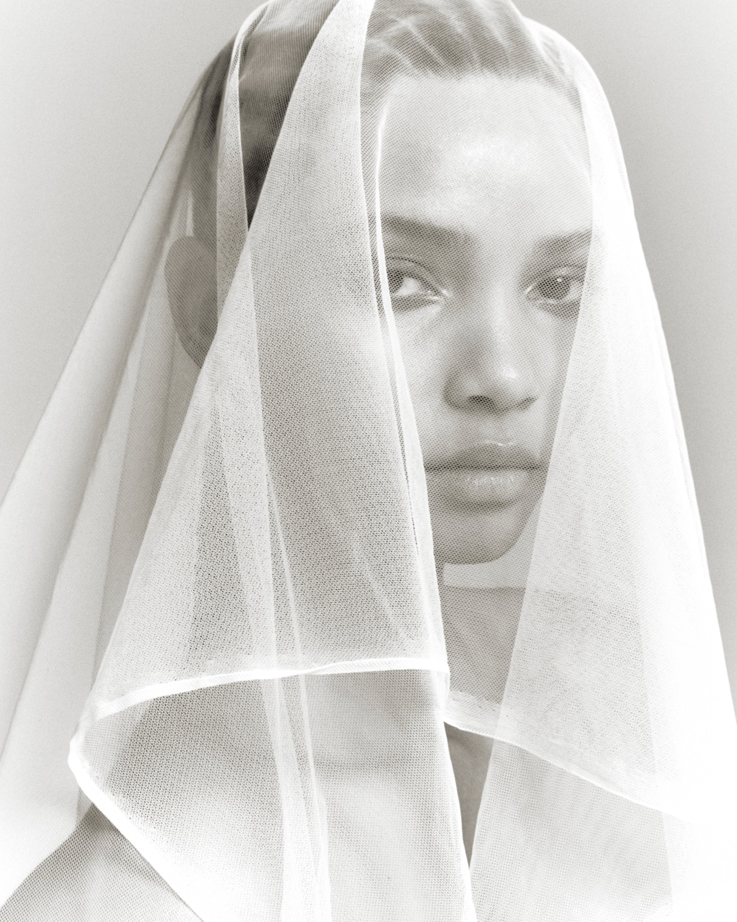 Monochrome portrait of an adult female with a neutral facial expression, draped in a sheer white veil against a plain studio backdrop.