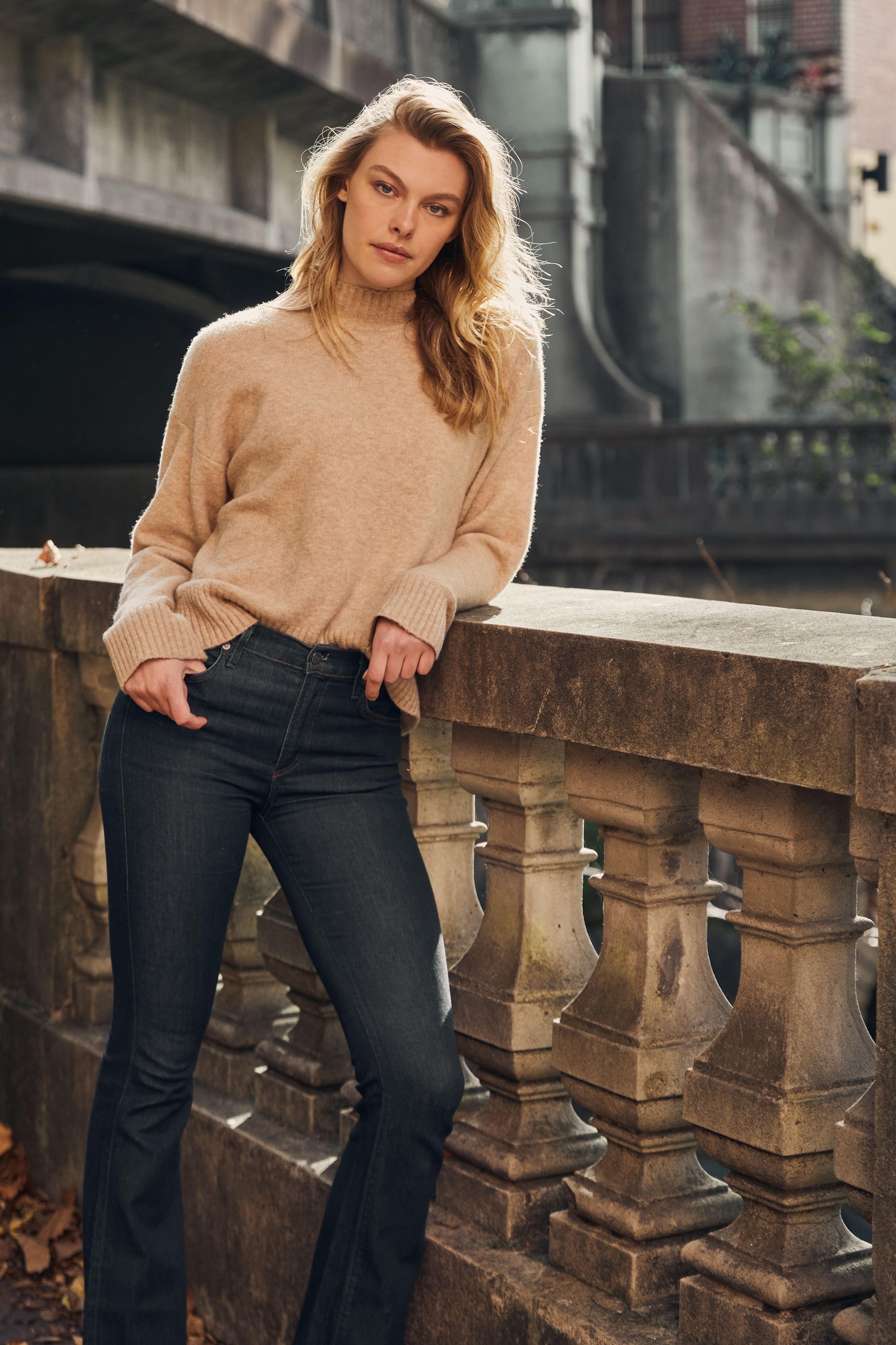 Woman in a beige knit sweater and dark blue flared jeans leaning against an ornate stone bridge railing