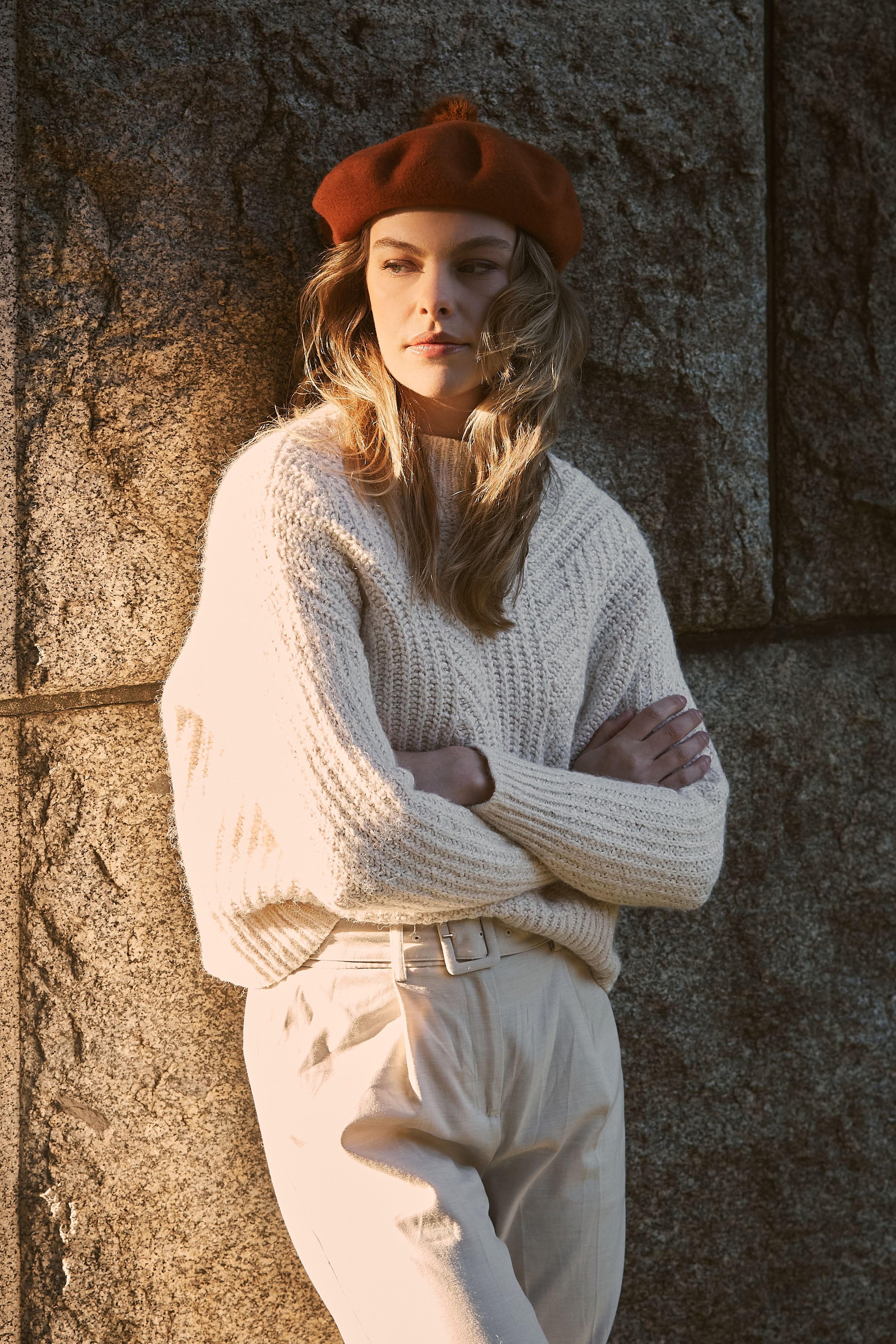 Portrait of a blonde adult female wearing a cream knit sweater, beige pants, and an orange beret, posing against a stone wall.