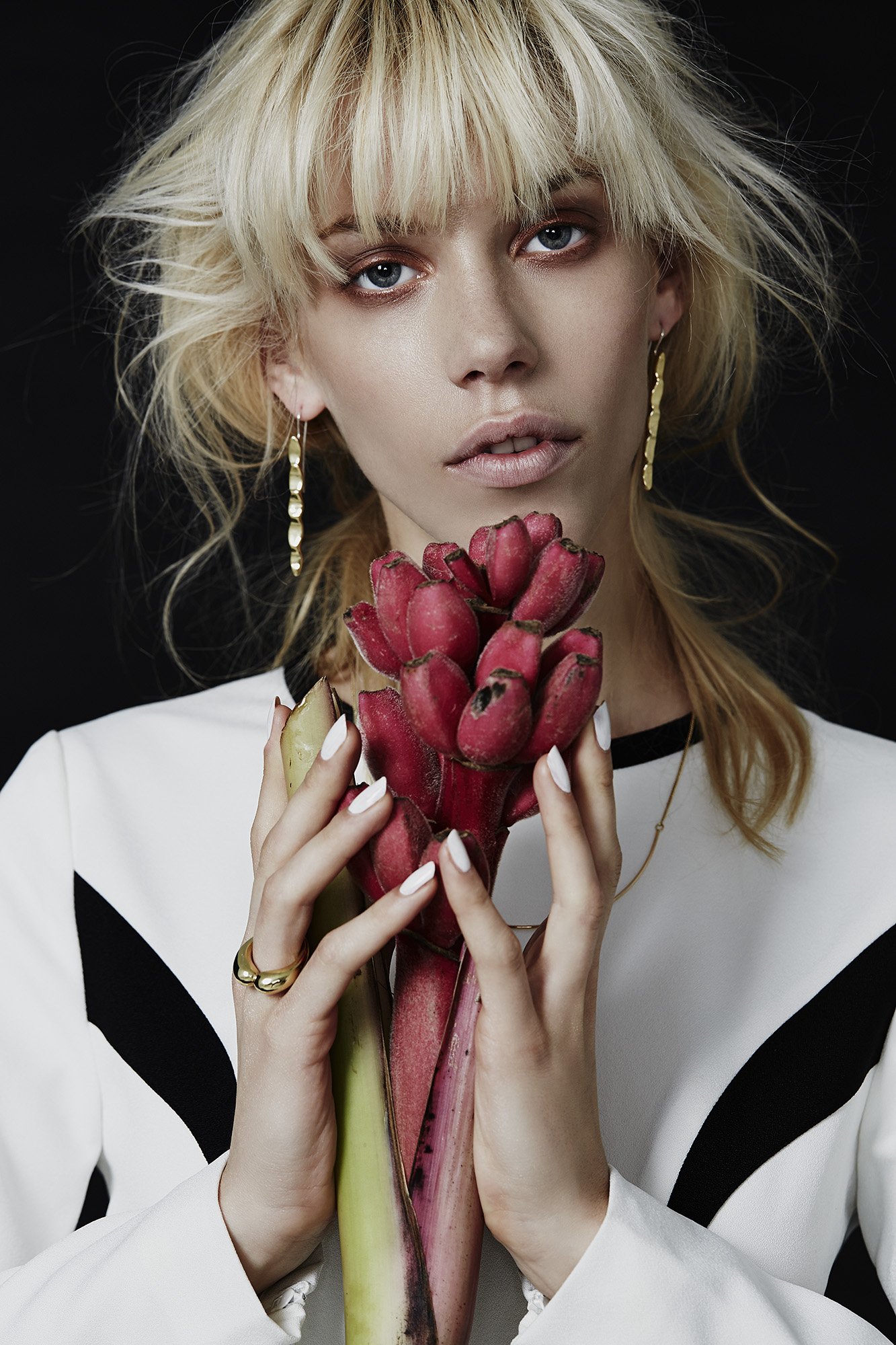 Close-up of a blonde woman holding a red torch ginger flower, wearing a white top and gold jewelry, against a black backdrop.