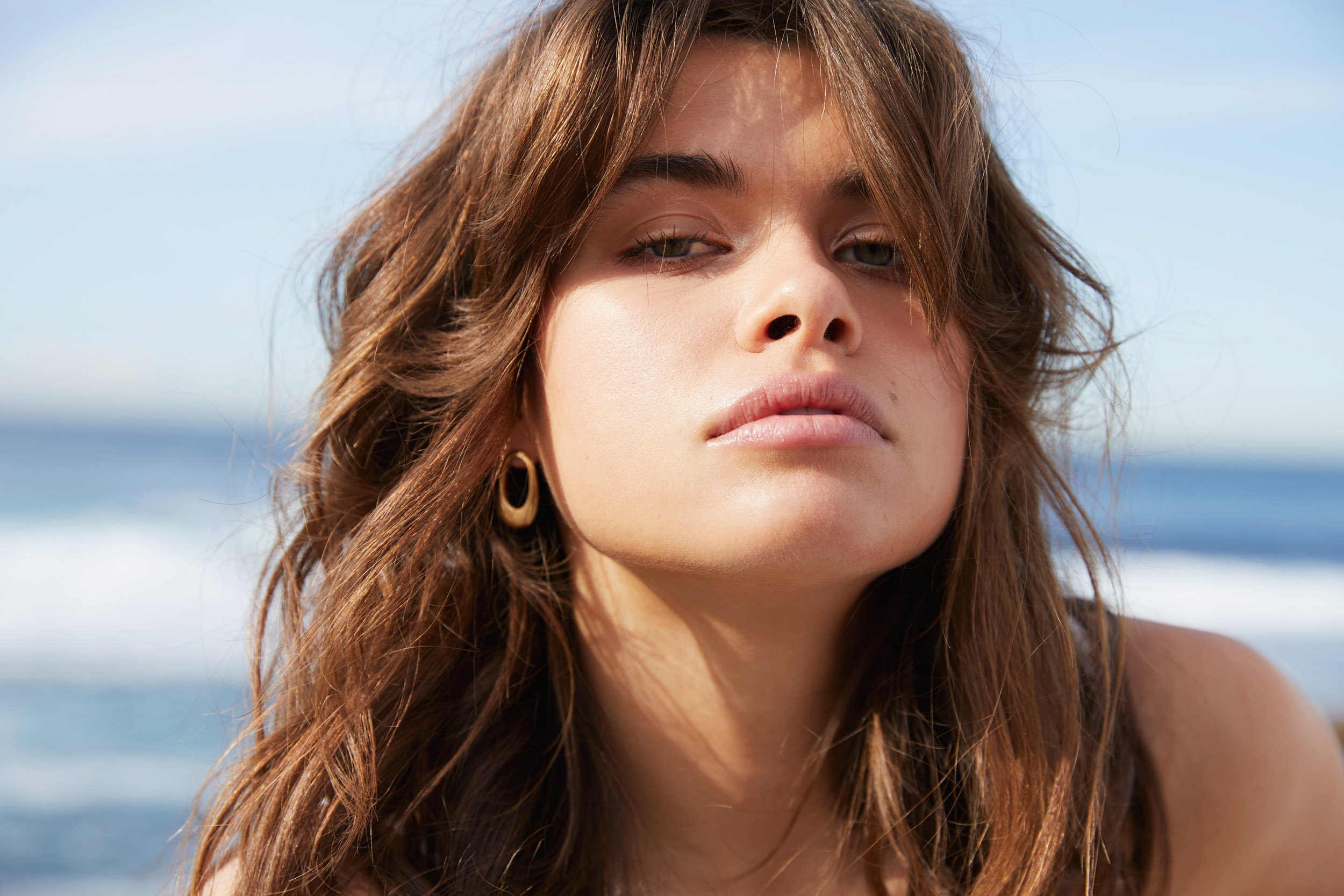 Close-up portrait of a young woman with brown wavy hair and gold earrings posing in an outdoor setting with a blurred background.