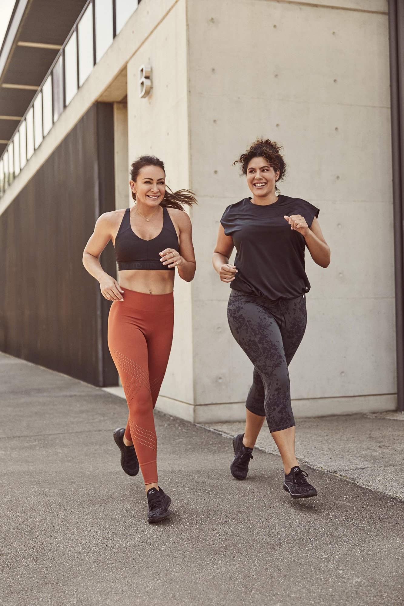 Two smiling women in athletic wear run outdoors on a paved surface near a modern concrete building in natural light. Fitness, exercise.